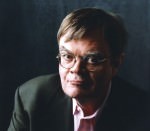 Garrison Keillor [Image description: headshot of Garrison Keillor, a white man with brown hair and glasses, wearing a pastel-colored button-down shirt and a black blazer, looking at the camera,]